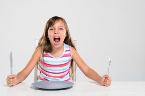 caucasian little girl sitting at a table with a fork and knife in her hand while screaming 