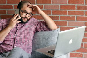 man sitting in front of a laptop with his hand on his head and phone to his ear.