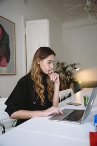 caucasian woman sitting at a desk looking at her laptop with her hand on her chin