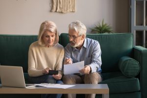 caucasian older couple sitting on a couch, the woman holding a calculator and the man holding a piece of paper.