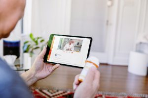 older man holding a tablet with a doctor on the tablet and holding medicine bottle in the other hand.