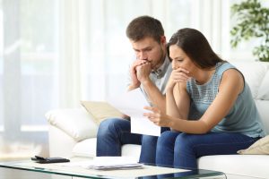 caucasian man and woman sitting on a couch looking worried with a piece of paper in the womans hand