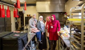 women in hijabs standing next to each other in a kitchen, with one woman sitting on the table smiling.