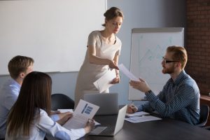 caucasian woman standing and handing a caucasian man a piece of paper in an office room setting with other people sitting at the desk