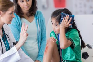 little girl with ice bag on her head and scratch on her shin with a doctor pointing 4 fingers up towards her