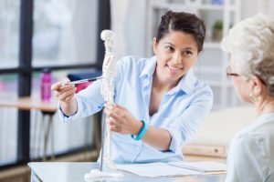 woman in a white labcoat pointing at a miniature skeleton's spine on her desk with an older caucasian woman sitting across from her