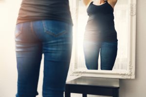 back of a thin woman with a reflection in the mirror of a bigger body.