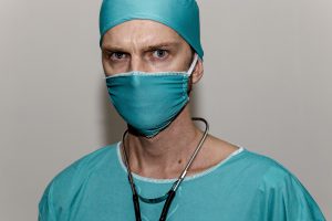 caucasian man that appears to look mad with teal scrubs on, mask and hat, as well as a stethoscope around his neck.