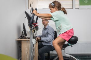 caucasian little girl on a stationary bike with wires connected and a man sitting down in the background watching
