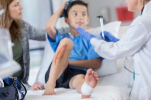 little caucasian boy laying down on a hospital bed with blood on his knee and gauze on foot with doctor checking him