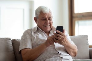 older caucasian man sitting on his couch holding a cell phone up smiling
