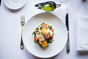 a white plate with a portion of salmon and vegetables with a fork on one side of the table and a knife on the other side.