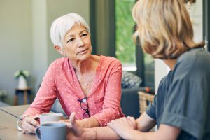 two older women talking while sitting at a table with coffee mugs on it
