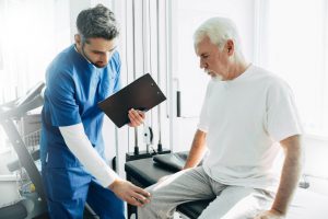 older man sitting on a table with a male doctor touching his knee with one hand and holding a clipboard in another.