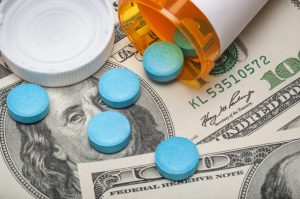 round blue pills spilling out of prescription bottle on top of hundred dollar bills.