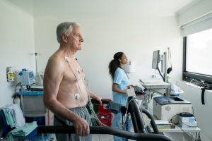 older caucasian man on a treadmill with no shirt on and wires stuck to his chest with a doctor in the background