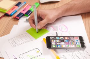 person's hand writing down on post it note with cell phone close by and a drawing of phone on paper with other writing surrounding it