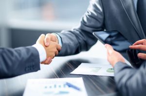 two hands in suits shaking over a table with paperwork on it.