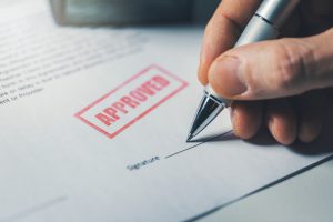 approved written on a paper with a person's hand holding a pen about to sign underneath the approved stamp