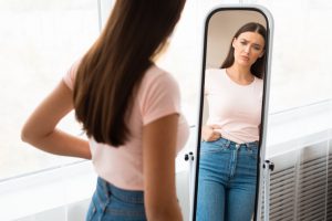 young teen girl in the mirror looking sad while pinching her stomach.