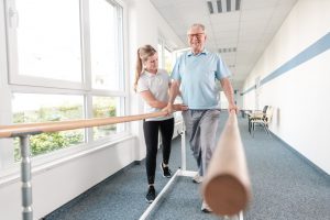 older Caucasian man holding onto a railing on both hands walking with the help of a younger caucasian woman.