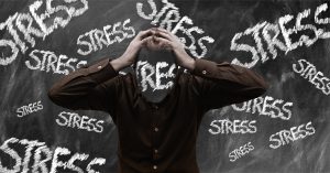 man with his hands on his head with the word stress on his face and stress written on a blackboard behind him.