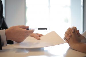 hand with a pen in it offering it to another hand with papers in the other hand.