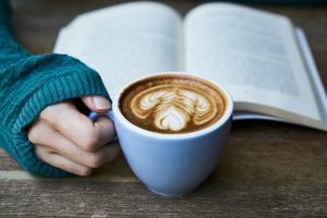 a caucasian hand holding a coffee mug with coffee in it and an open book behind the cup.