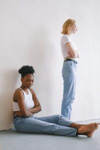 caucasian woman standing with her back to the young african american woman sitting on the floor looking away from her with her hands crossed.