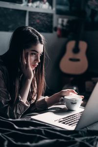 young caucasian woman sitting down in front of a computer at night with coffee in her hand.