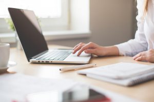 caucasian womans hand on the keyboard of her laptop with papers and a pencil next to it.