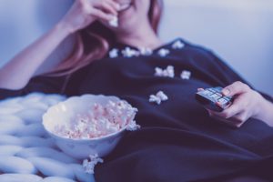 caucasian woman sitting down with a remote in one hand and the other with popcorn from a bowl next to her.