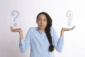 woman in a blue dress shirt shrugging her shoulders with a question mark on each hand