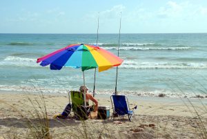 man sitting on a beach under a rainbow umbrella with fishing pole in the sand