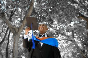 african american young man in a black gown holding his black graduation cap while smiling