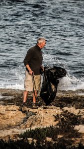 older man picking up trash along the beach while holding a black garbage bag