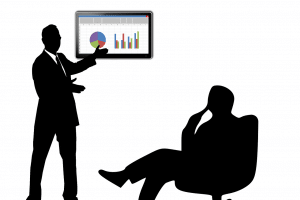 person sitting down and another person standing up pointing towards a screen with graphs on it