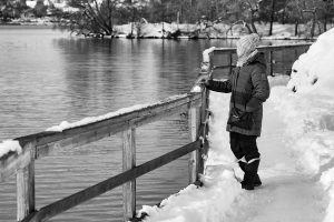 black and white picture of a woman looking out at water while bundled up and snow on the ground