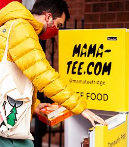 person in a yellow puffer jacket holding food items with the yellow mama-tee fridge open