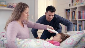 parents holding a thermometer over a young girls head who is in bed