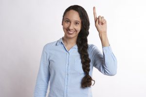 woman with brown hair and light blue shirt standing in front of a white wall with a hand with index finger raised